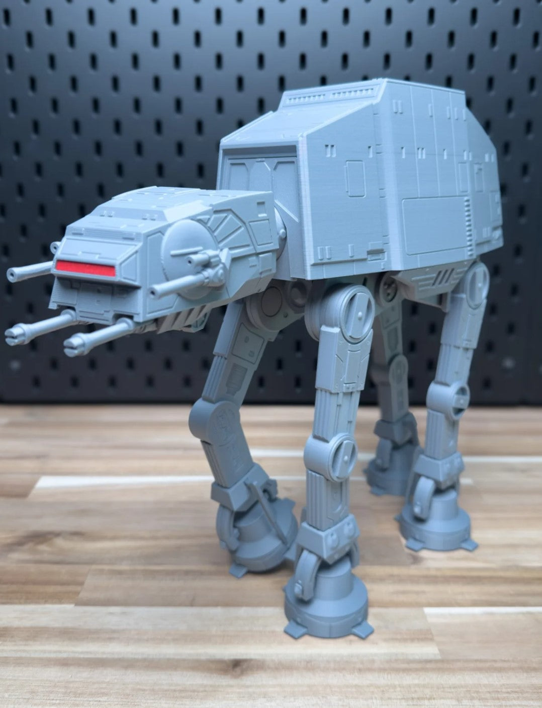 Gray toy AT-AT walker model on a wooden surface with a black pegboard background