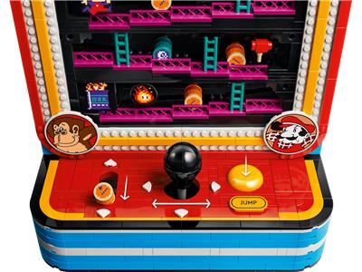 A toy arcade machine built from interlocking blocks, featuring a red control panel with a black joystick, yellow buttons, colorful game screens, and cartoon character medallions on a white background.