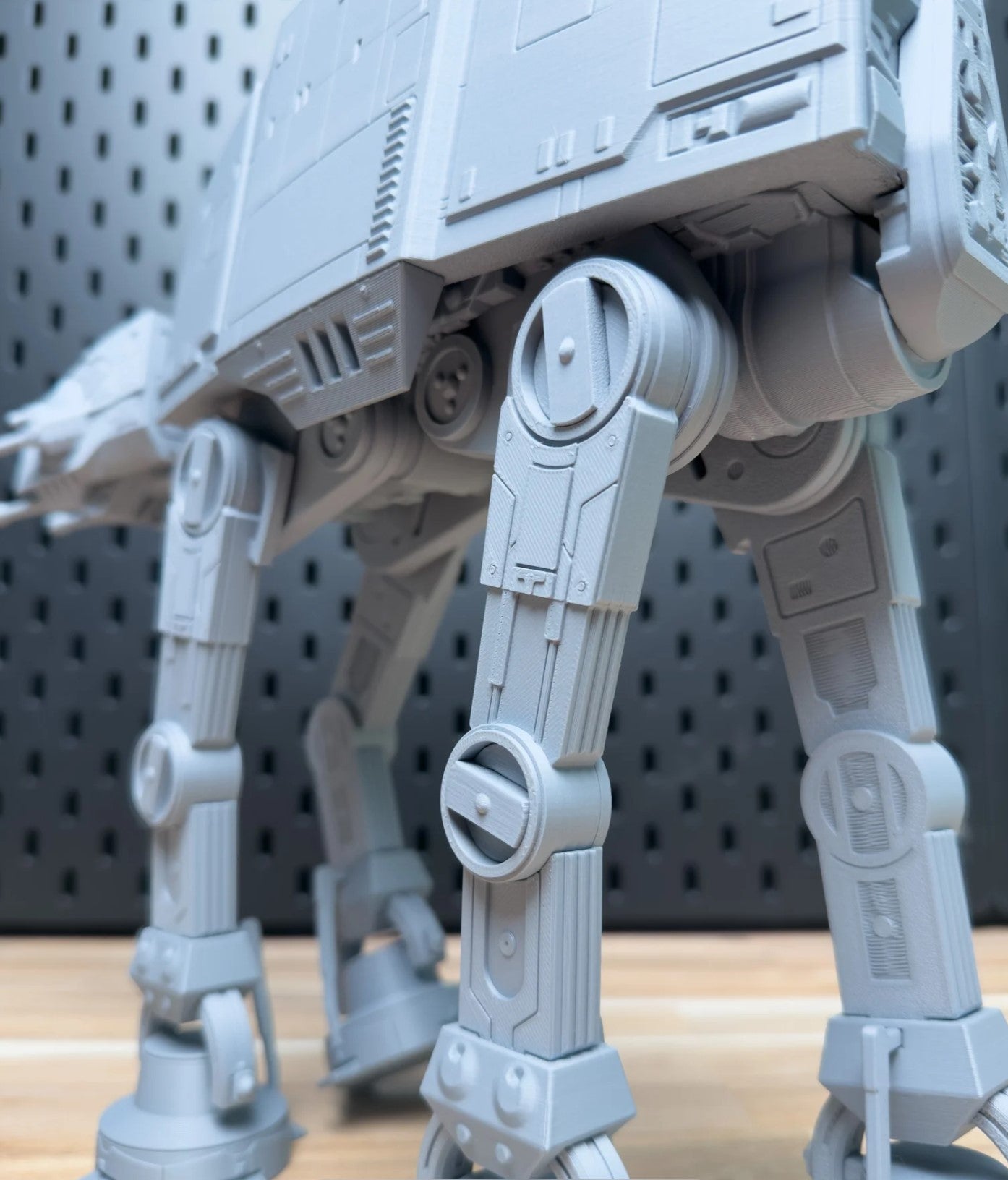 Close-up of a gray toy AT-AT walker model on a wooden surface with a gray pegboard background.